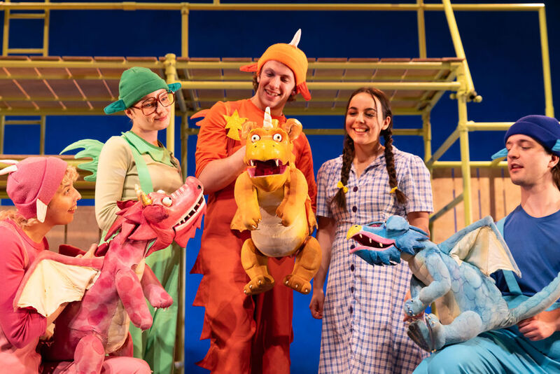 Five smiling actors from the Zog theatre production stand together on stage, happily interacting with colourful pink, orange, and blue dragon puppets.