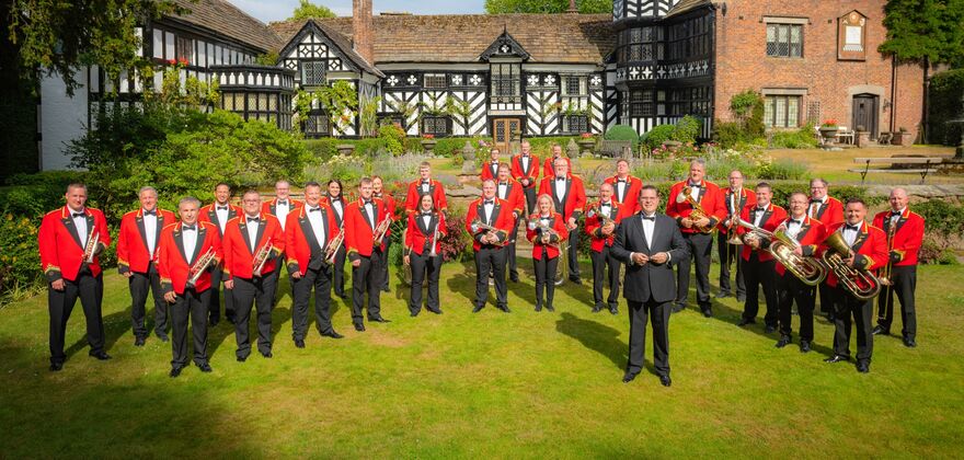 A large group portrait of Foden's Band standing in front of a historic black-and-white half-timbered building.