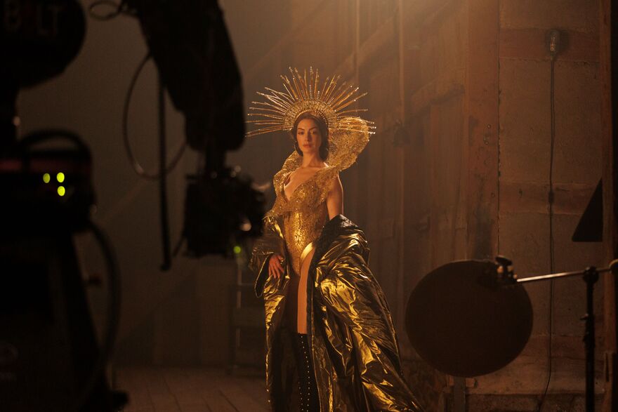 A woman stands on a sound stage. She is dressed in a gold body suit, gold coat and crown. 