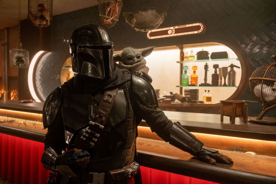 The Mandalorian (a person in armour with a helmet) and Grogu (a green alien like character, similar to Yoda from Star Wars) in a bar. 