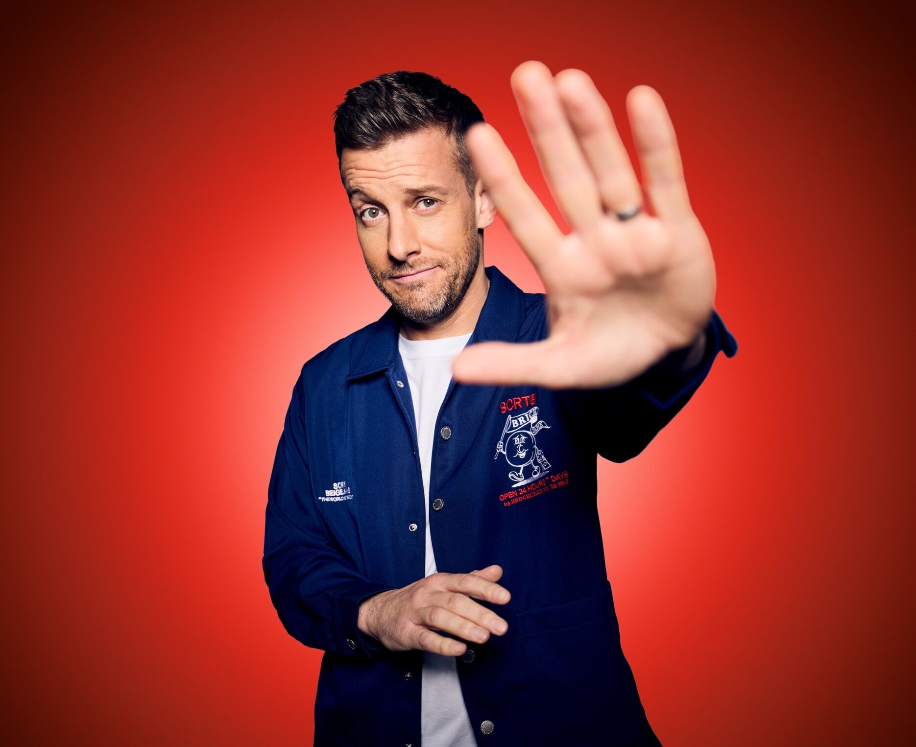 Chris Ramsey in a blue jacket with one hand up with a red background