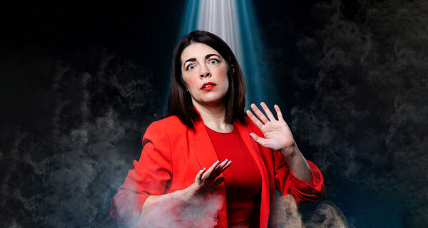Comedian Rosie Holt in a bright red suit looks surprised with her hands raised, standing in swirling smoke under a bright spotlight.