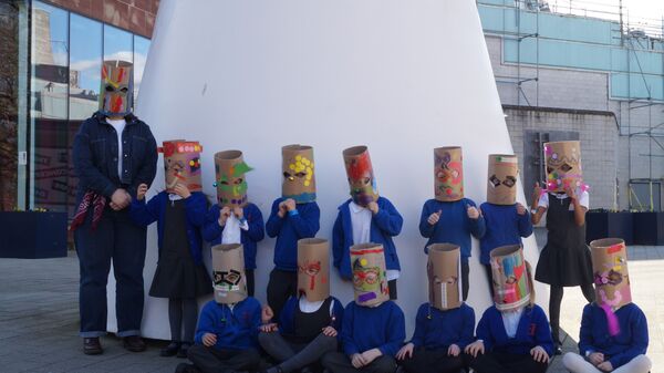 Children and a teacher stood in front of a large white sculpture with masks on 