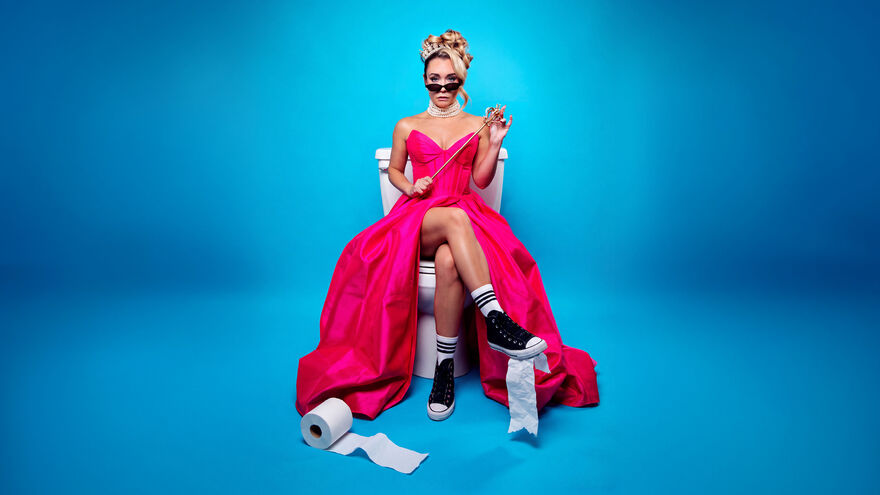 Sophie McCartney in a long pink dress and sunglasses sat on a toilet