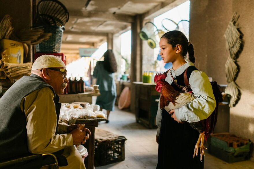 A young girl holds a cockerel as she speaks to an older man in a market. 