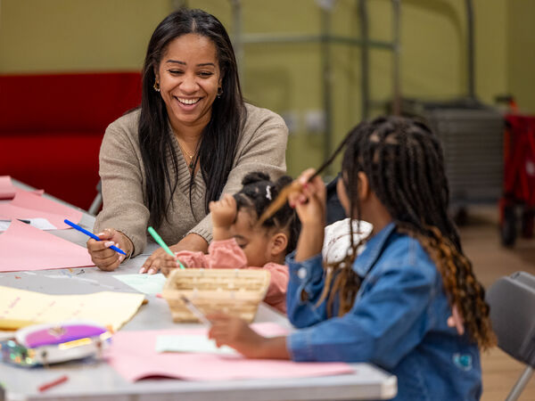 A woman sits at a workshop table with two young children who are busy drawing. A woman sits at a workshop table with two young children who are busy drawing.