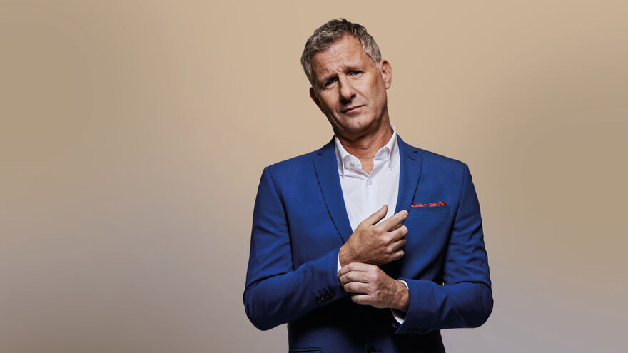 Adam Hills in a blue suit adjusting his cuff