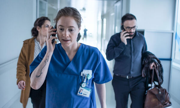 A nurse talks on the phone as she walks down a hospital corridor. A man and woman walk behind her. 