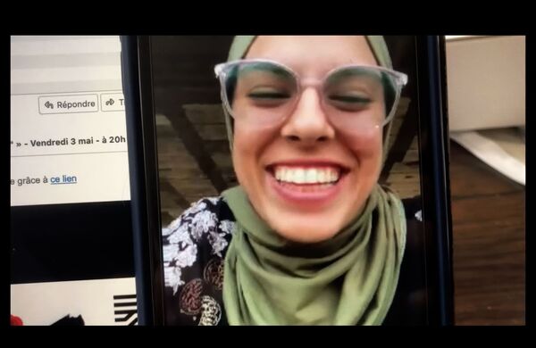 A young woman in glasses and a head covering smiles through a mobile phone screen. 
