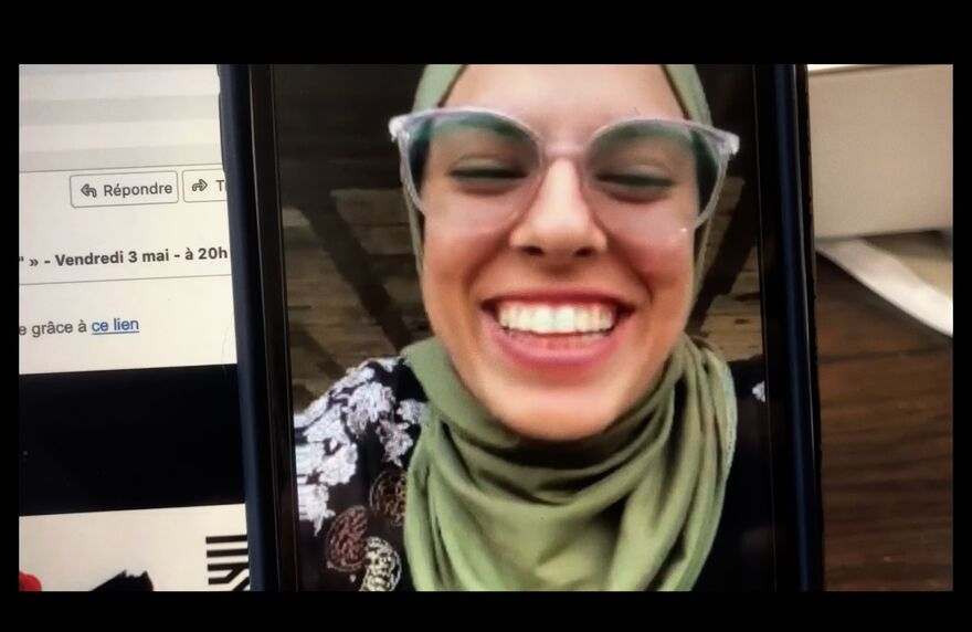 A young woman in glasses and a head covering smiles through a mobile phone screen.