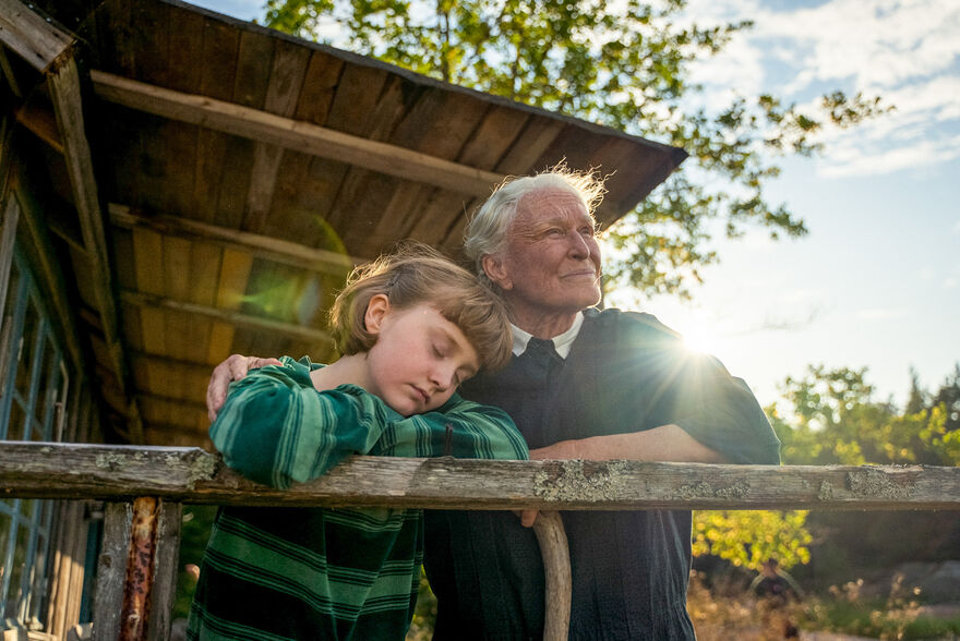 A young girl and her grandmother stand together on an outdoor porch. 