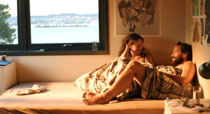 A couple lie in bed together covered in a duvet.