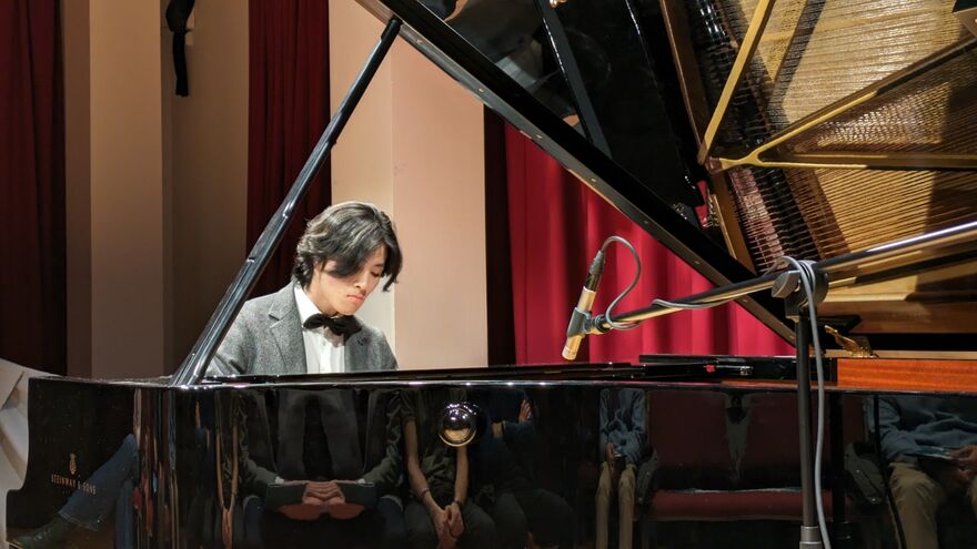 A young man in a grey blazer plays a grand piano on stage.