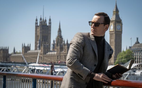 A man in suit and sunglasses leans on a railing, holding a book. The Houses of Parliament are in the background. 