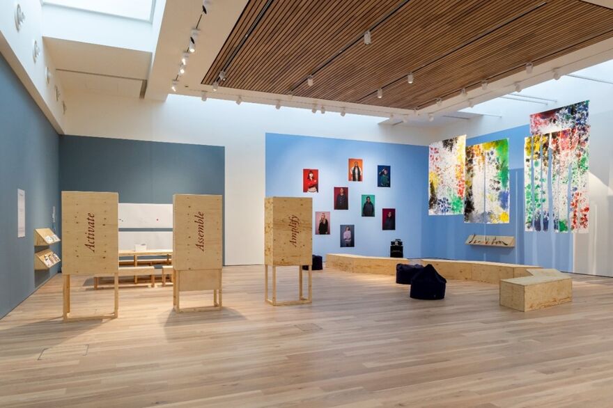 Interior view of an art gallery with walls displaying various colorful paintings and photographs. In the center, there are several wooden installations and seating areas for viewers.