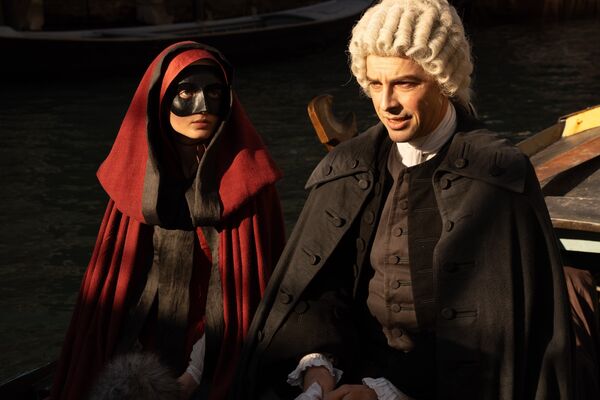 A woman in red cloak and a black masquerade mask sits next to a man in white wig and all black dress on a venetian gondola.  