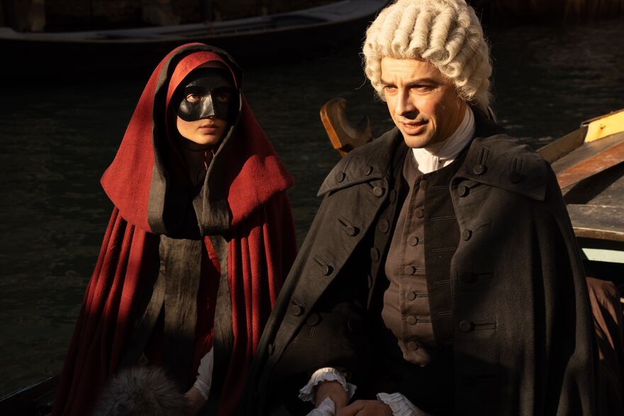 A woman in red cloak and a black masquerade mask sits next to a man in white wig and all black dress on a venetian gondola.  