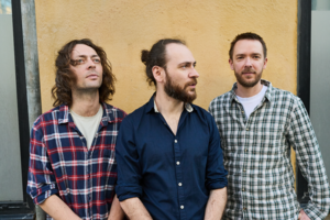 3 men stood outside in front of a yellow wall