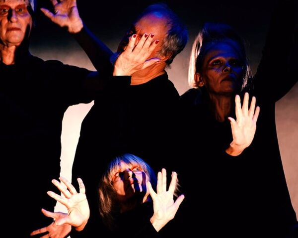 Four older dancers in black tops performing on a dark stage, their hands raised with palms facing outwards under dramatic blue and orange lighting.