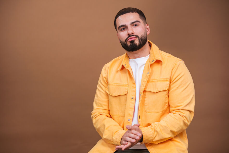 A studio portrait of comedian Kae Kurd sitting against a plain brown background.