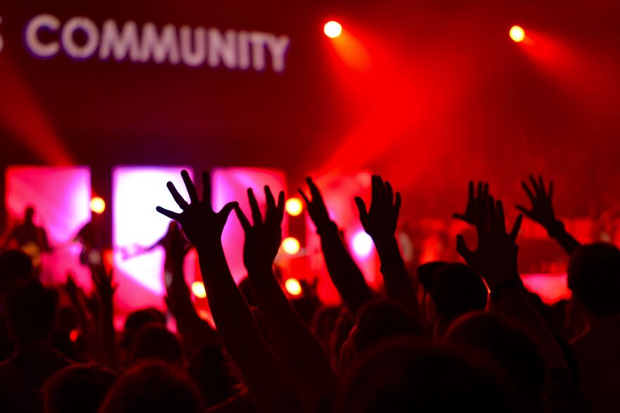 People's arms in the air at a concert with the word community above the stage