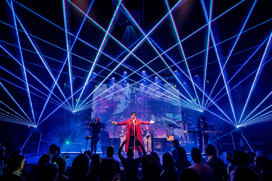 A singer in a red coat leads The 80s Show performance under a spectacular display of crossing blue laser beams.