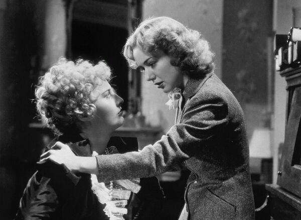 A black and white image of two women looking at each other. One is stood against a piano, the other sat on a stool. A black and white image of two women looking at each other. One is stood against a piano, the other sat on a stool.
