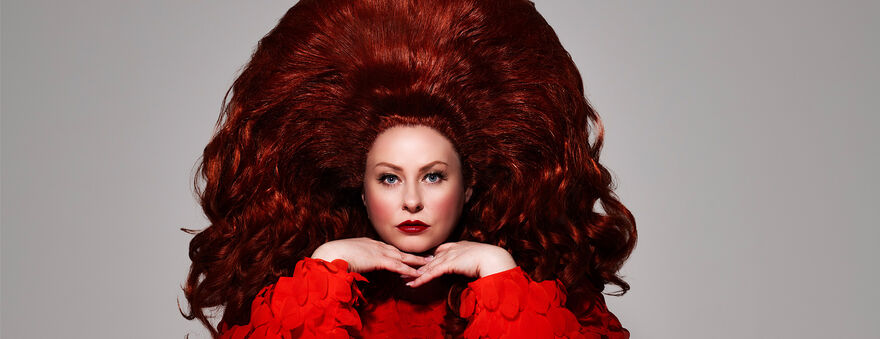 Amy Gledhill with both hands under chin in a red dress and big red hair