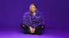Joanne McNally wearing large purple ruffled top, sitting cross-legged in front of a purple background