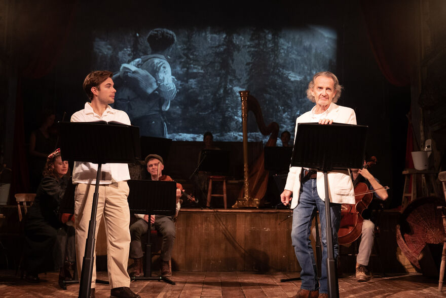 Anton Lesser and Charlie Hamblett are at the front of the stage with stands in front of them. Behind them, a small orchestra, harp and an image of a soldier with his back to the audience are seen