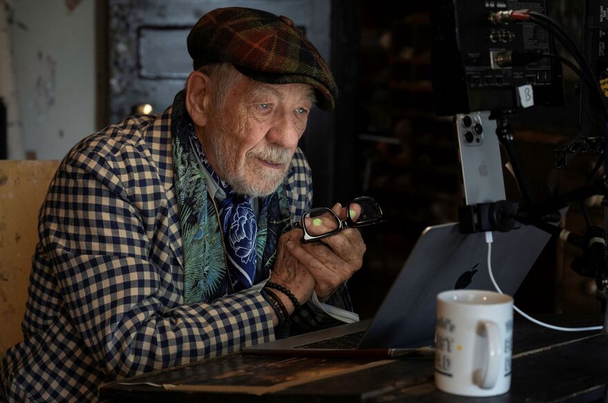 An older man wears flat cap and checked shirt as he looks at his phone and laptop. 