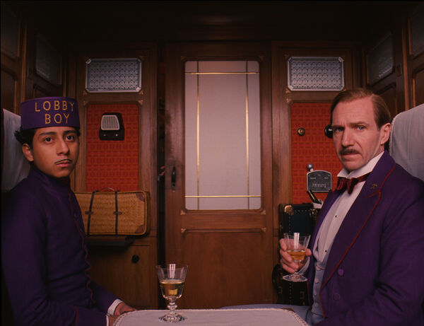 A hotel concierge and lobby boy, both dressed in purple, sit opposite each other in a train carriage looking out of the window. 