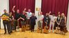 A group of smiling musicians holding guitars, a violin, and a ukulele, waving at the camera in front of red stage curtains.