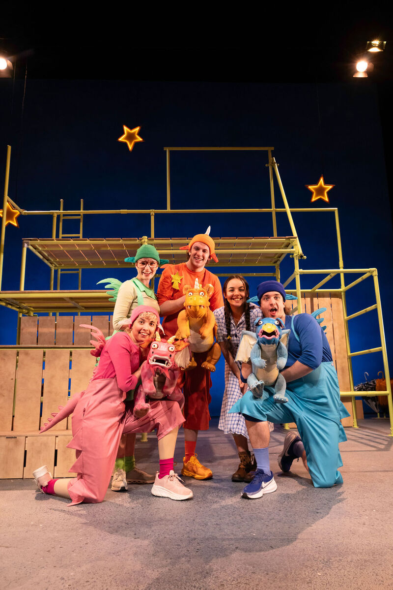 Five smiling actors from the Zog theatre production pose together on stage in colourful costumes, holding pink, orange, and blue dragon puppets.