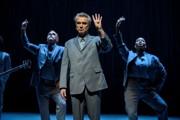 David Byrne holds up four fingers. Two dancers pose behind him with their arms up. David Byrne holds up four fingers. Two dancers pose behind him with their arms up.