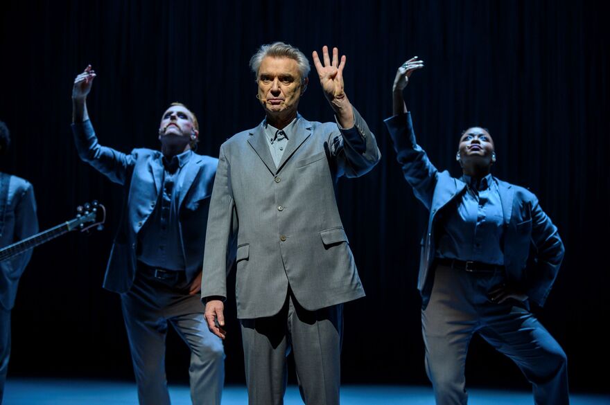David Byrne holds up four fingers. Two dancers pose behind him with their arms up. 