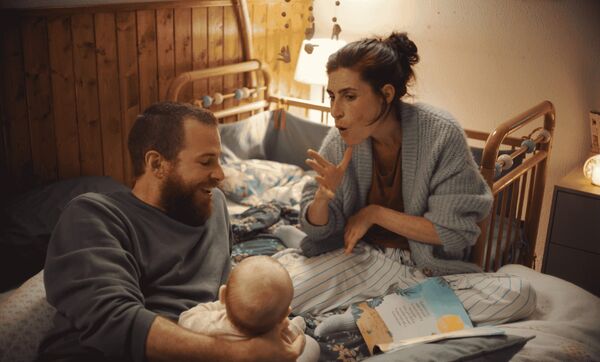 A couple sits on a bed with a baby. The woman signs to the baby. 