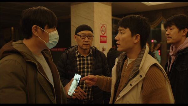 A group of men stand together. One is wearing a face mask whilst on a video call. 