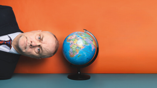 Jack Dee appearing from the side of the frame with his head next to a globe