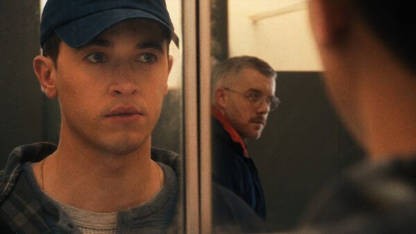 A young man looks into a mirror, meeting eyes with another man walking behind him. 