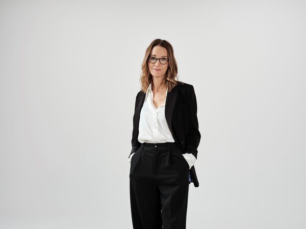Susie Dent in a black suit with a white blouse and glasses on a grey background