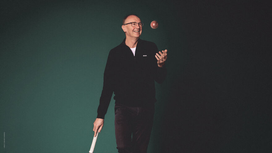 Jonathan Agnew leaning on a cricket bat trowing a cricket ball in the air