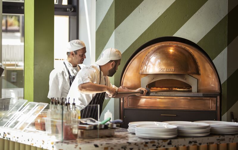 Two chefs taking pizzas out of the pizza oven in Benugo