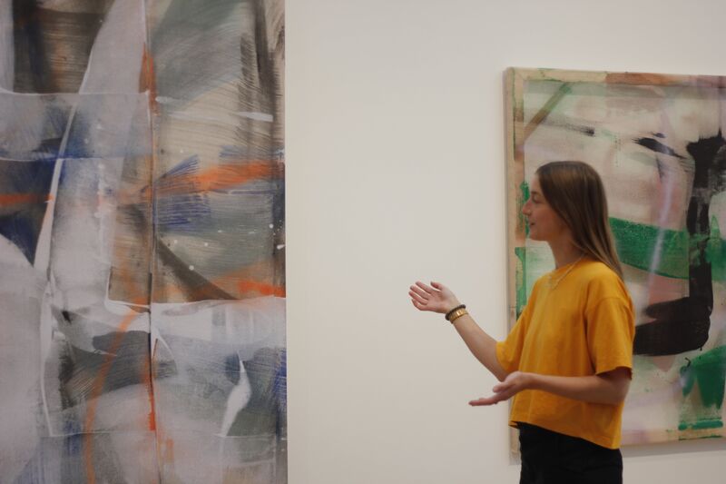 Woman in a yellow shirt gesturing towards an artwork on the wall 