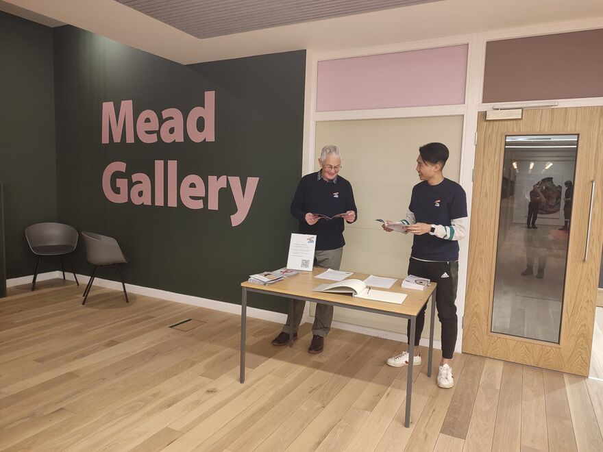 Two male volunteers standing behind a desk in front of the Mead Gallery