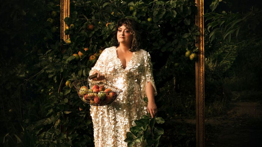 Comedian Kiri Pritchard-McLean in a sequined gown holding a basket of apples, framed by a golden picture frame in an orchard.