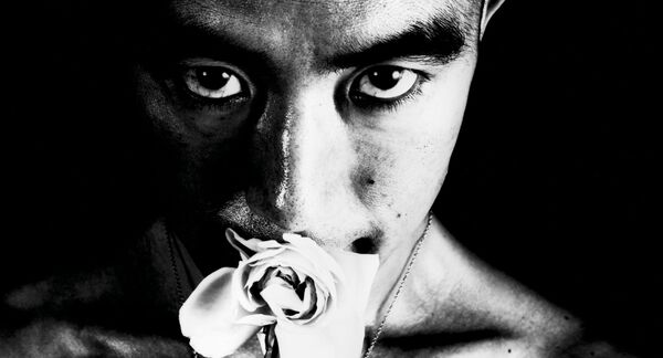 A black and white, close-up image of a man smelling a rose. He looks straight to camera. 
