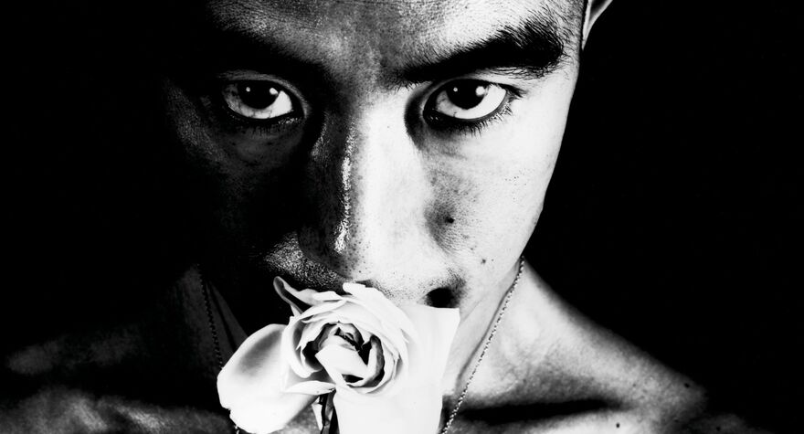 A black and white, close-up image of a man smelling a rose. He looks straight to camera.
