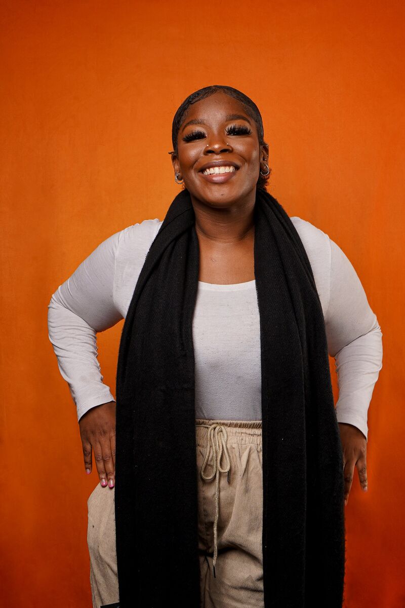 Laaiqah wearing a long black scarf standing in front of an orange background