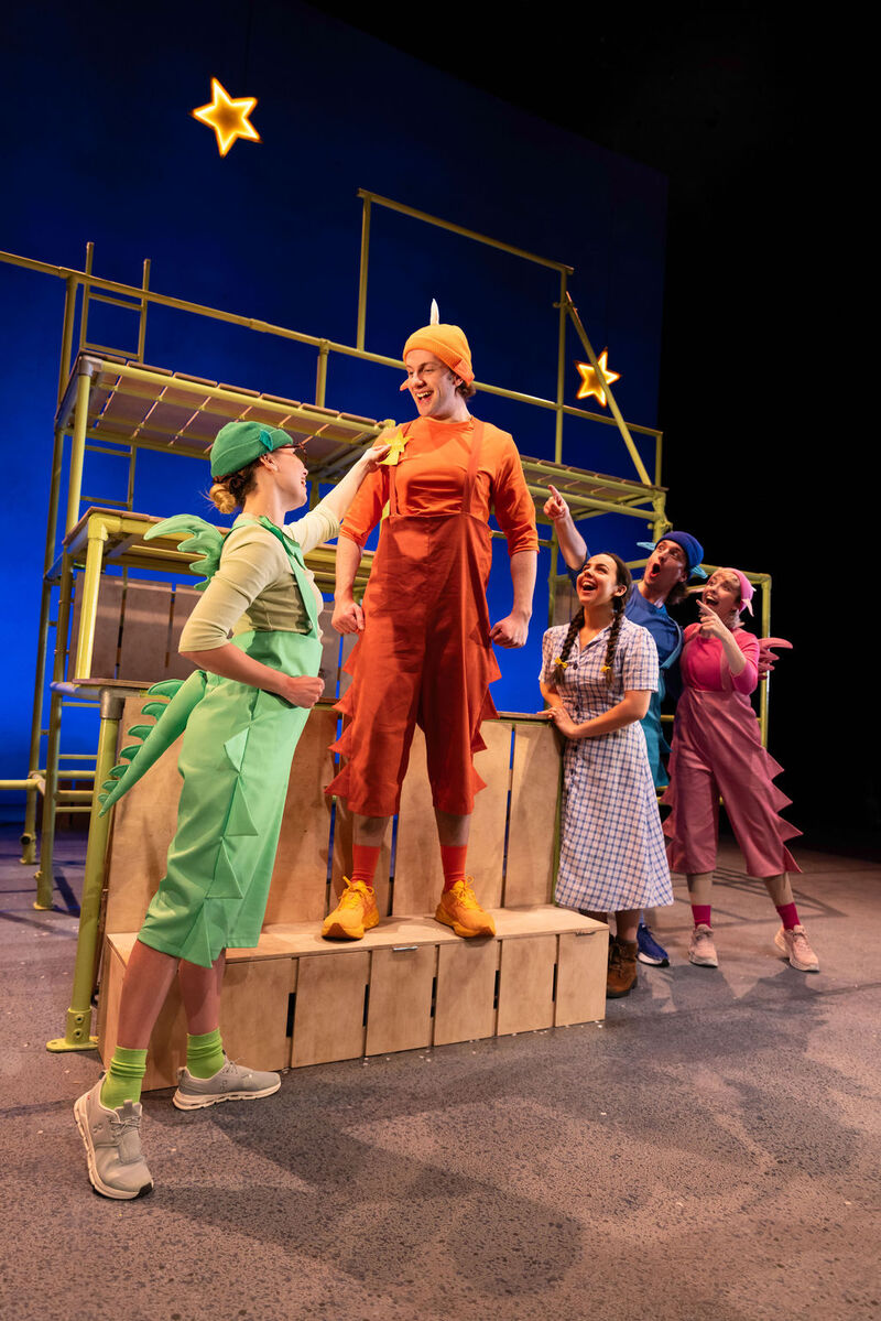 Five actors from the Zog theatre production gather on a scaffolding stage set as one excitedly pins a gold star onto the chest of a smiling actor in an orange dragon costume.
